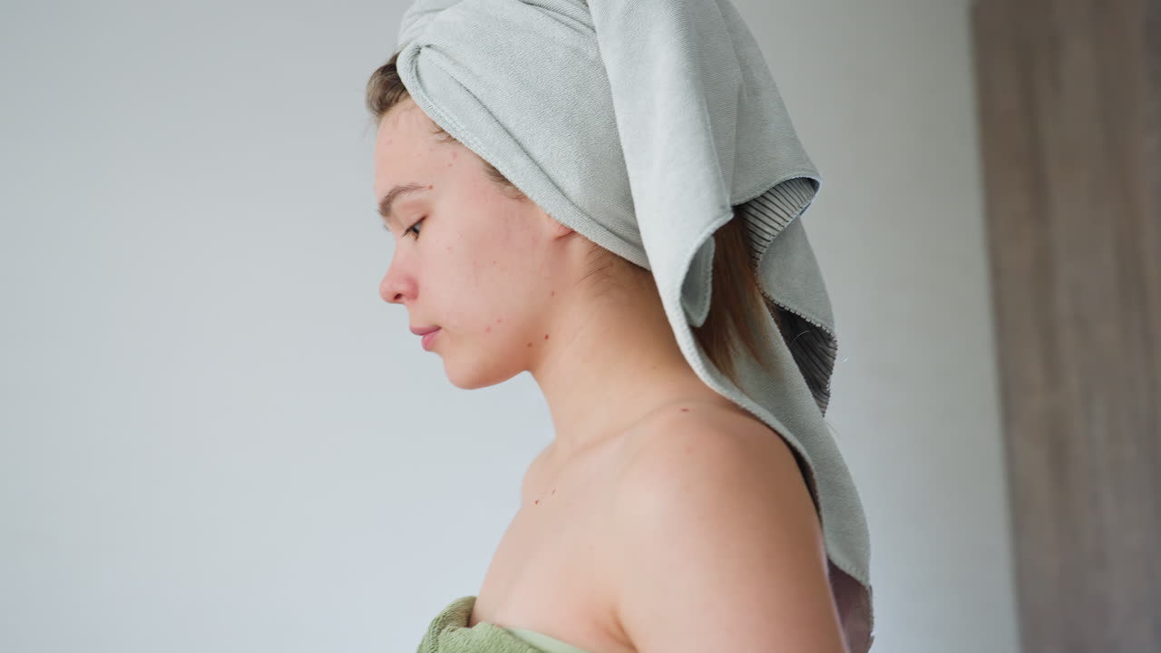 close up view of woman wrapped in green towel adjusting grey head towel while seated on sofa in bright living room with sheer curtains chandelier overhead capturing post-shower self-care moment