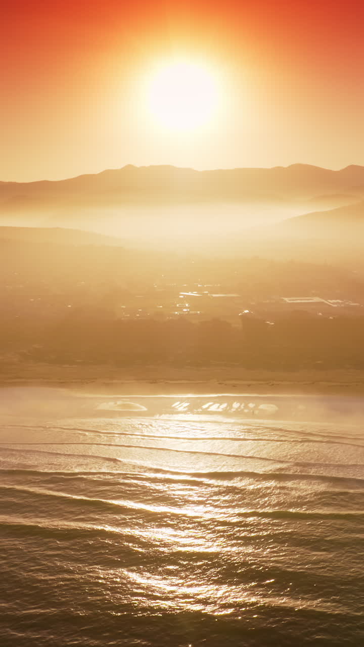 Sun reflecting in the sea water near the shore. Silhouettes of mountains and some features of city hardly seen in the rays of sunset. Vertical video