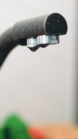 drops of clear water drip from dark modern faucet into sink. leaking plumbing fixture in stylish black and white kitchen on blurred background macro