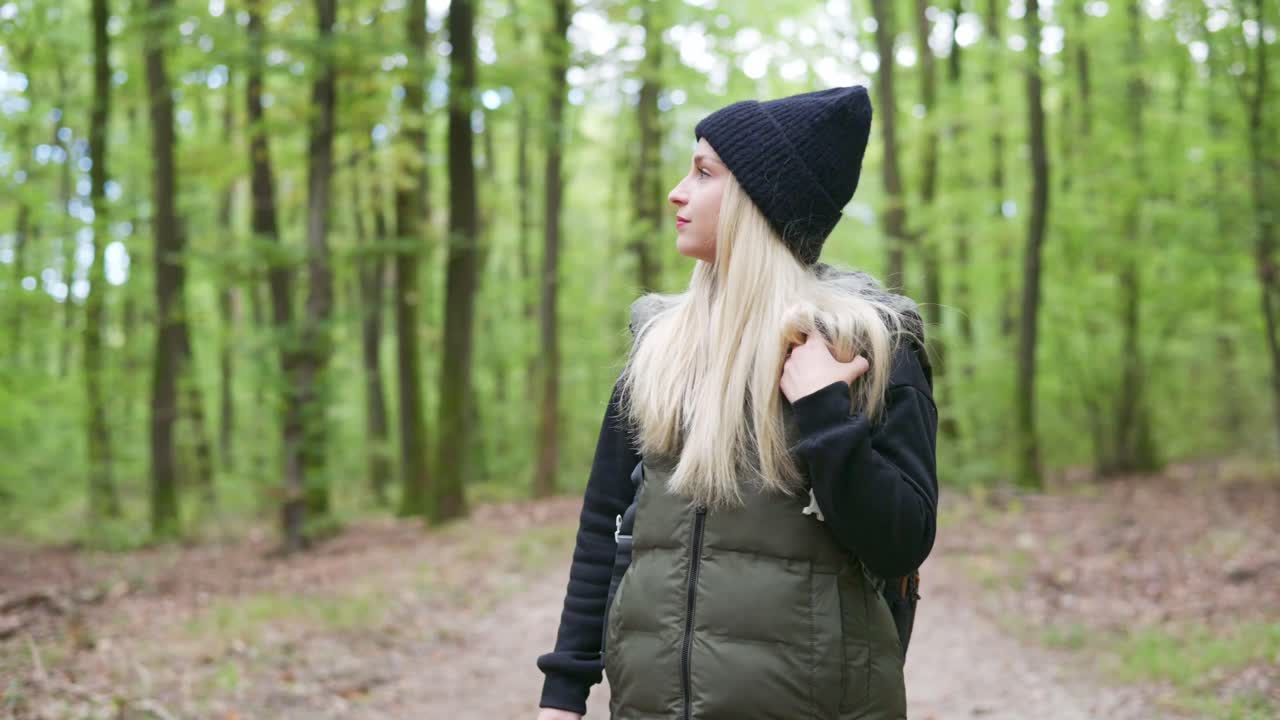 A woman with long blonde hair and a black beanie looks up while walking through a peaceful forest. She wears a warm vest and sweater, capturing the calm and reflective mood of an autumn day in nature