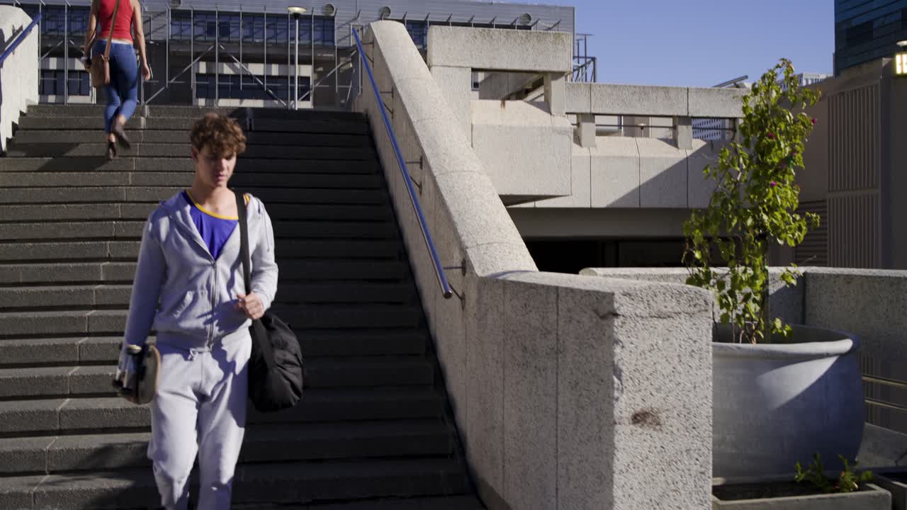 Coppia che si avvicina ai gradini della piazza, donna che sale con il telefono in mano, uomo che scende con skateboard e borsa, la telecamera segue il movimento.