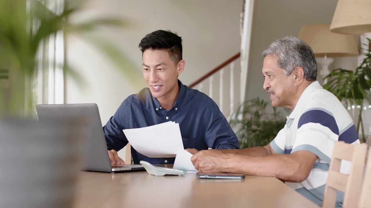 Diverse financial advisor and senior man discussing paperwork and using laptop at home, slow motion