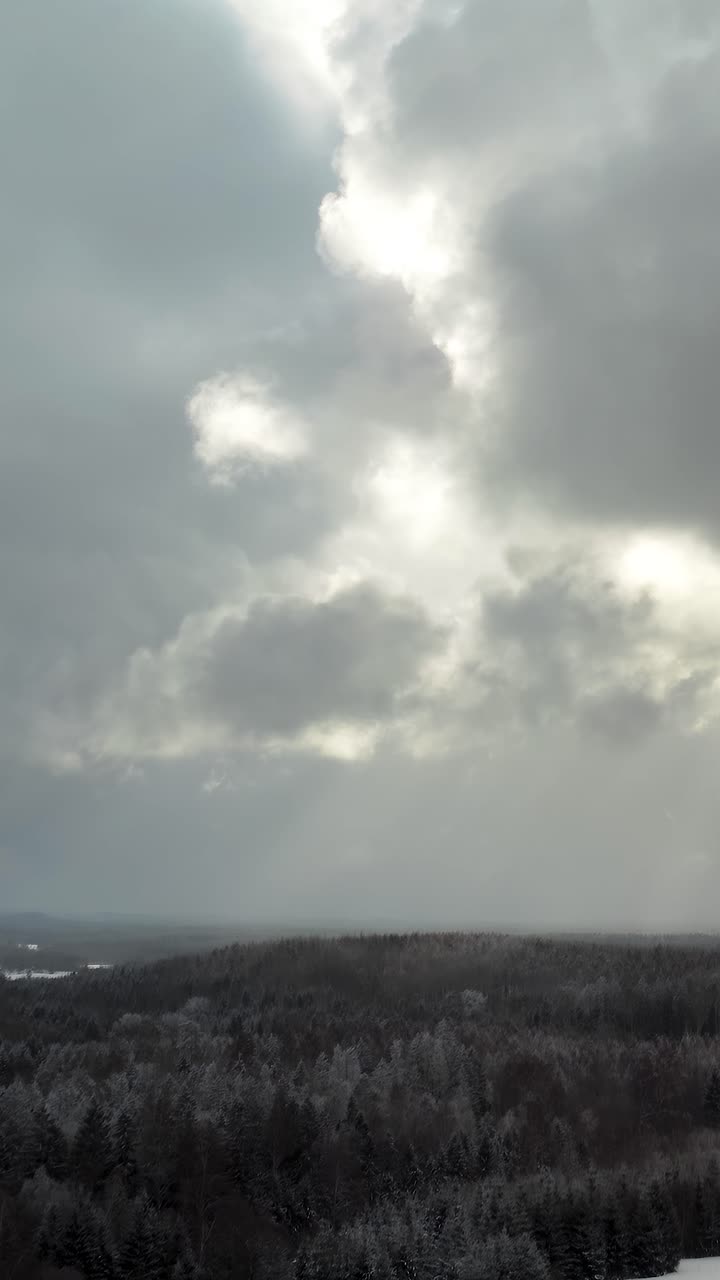 Vertical aerial view of thick clouds over the forest landscape in countryside. Snow covered ground and sun rays through clouds in winter. Snow storm approaching. Dramatic clouds.