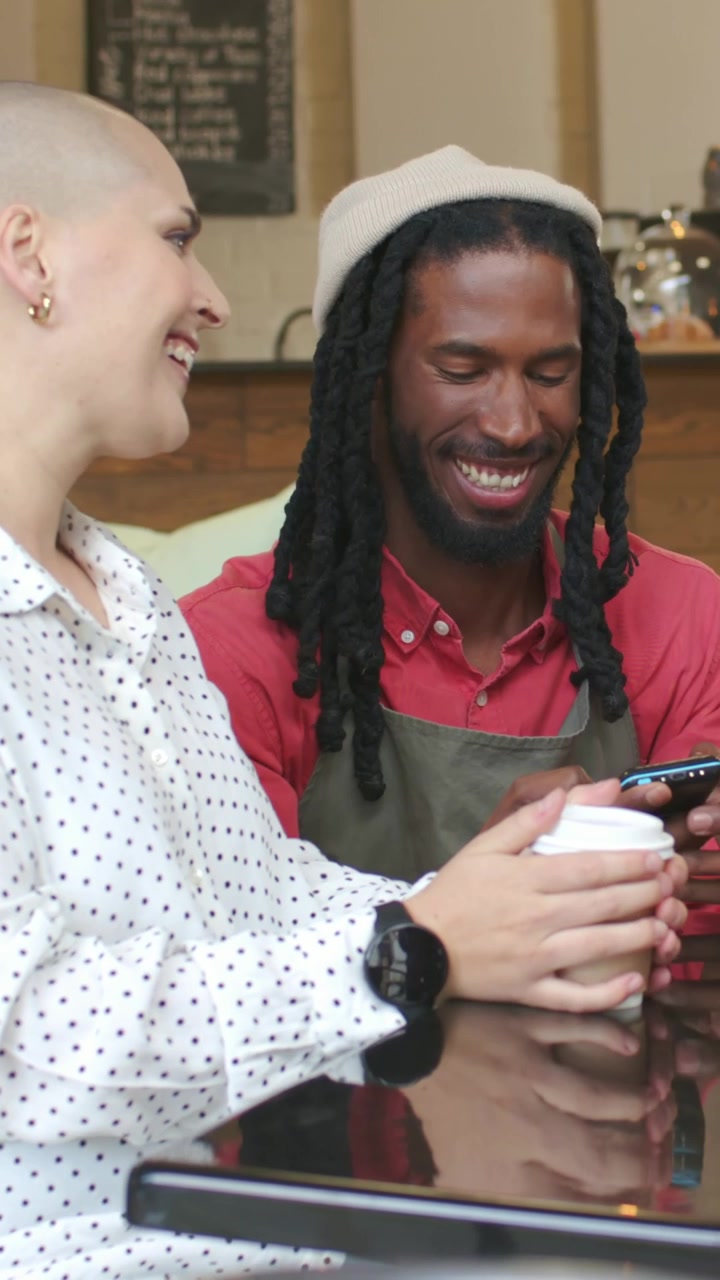 Vertical video: Barista in apron showing phone to woman with cup at cafe, then walking to counter
