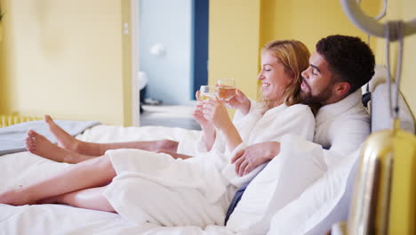 happy mixed race young adult couple reclining on the bed in a hotel room in bathrobes holding glasses of wine, close up