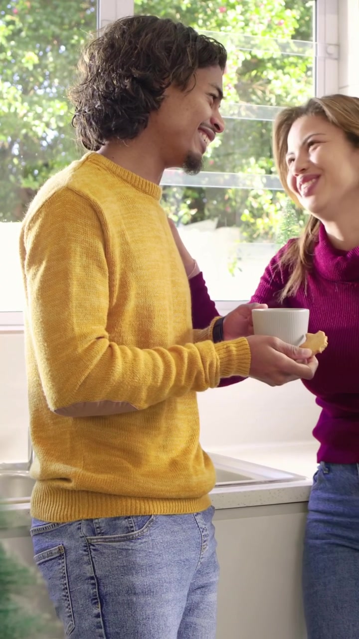 Video verticale: Uomo con maglia dorata che offre un cucchiaio, dando da assaggiare a una donna al bancone della cucina con una tazza.