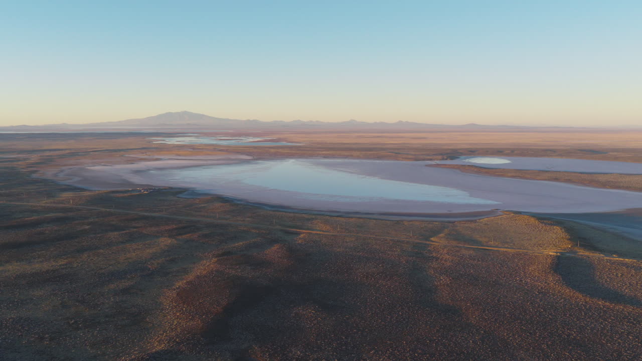 Aerial drone view of Salinas del Diamante basin in Mendoza, Argentina, reveals vast salt flats surrounded by arid terrain with the Andes in the distance under warm evening light