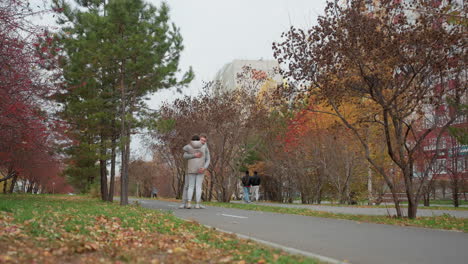 siblings dressed in identical outfits walk up to each other and share warm hug along tree-lined urban pathway as people stroll in background and colorful autumn branches sway gently in wind