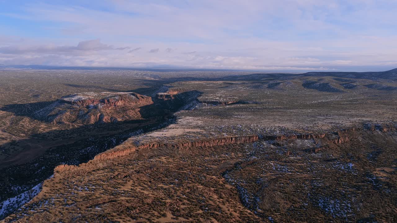 Una vista aerea mette in risalto l'ampio altopiano desertico e un prominente crinale roccioso con strati geologici visibili. Una leggera spolverata di neve invernale copre l'aspra natura selvaggia del sud-ovest.