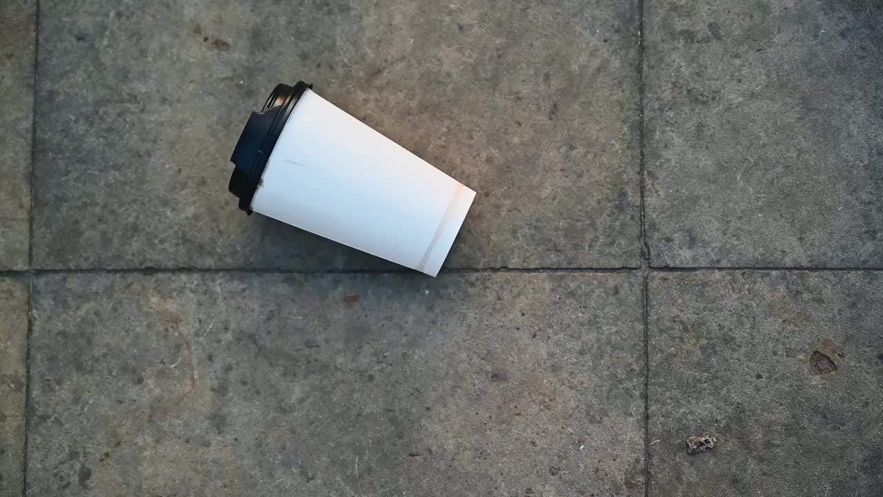 Man's feet hits an empty paper cup of coffee from the pavement at sunlight. Ecology idea. Slow motion. Top view