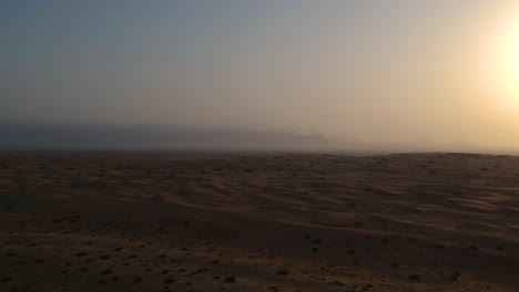 Drohne-Fliegt-Während-Des-Sonnenuntergangs-Durch-Die-Wüste,-Während-Sich-Sanddünen-Mit-Bergen-Im-Hintergrund-In-Wahiba-Sands-Im-Oman-Auf-Den-Betrachter-Zubewegen