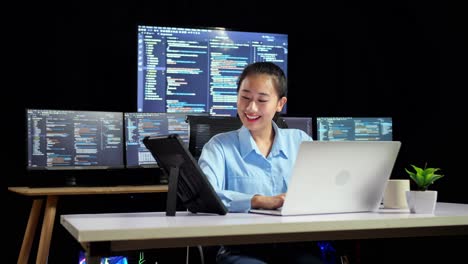 asian female programmer looking at tablet and writing code by a laptop using multiple monitors showing database on terminal window desktops in the office