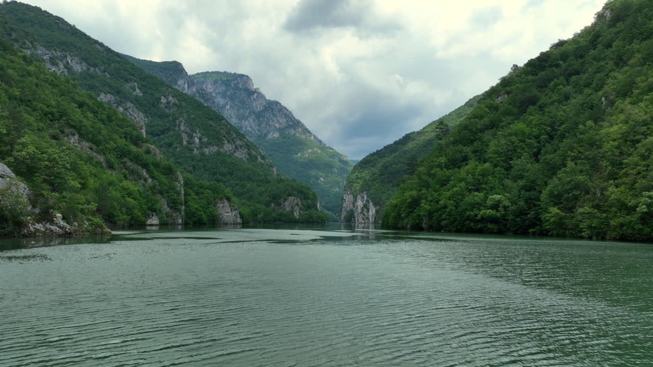 Pov from a boat cruising along a serene river surrounded by lush green mountains under a cloudy sky