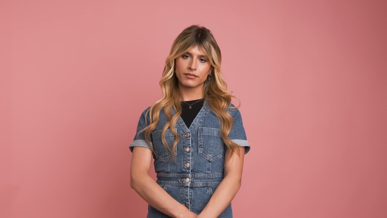 Confident transgender woman in denim dress against pink background