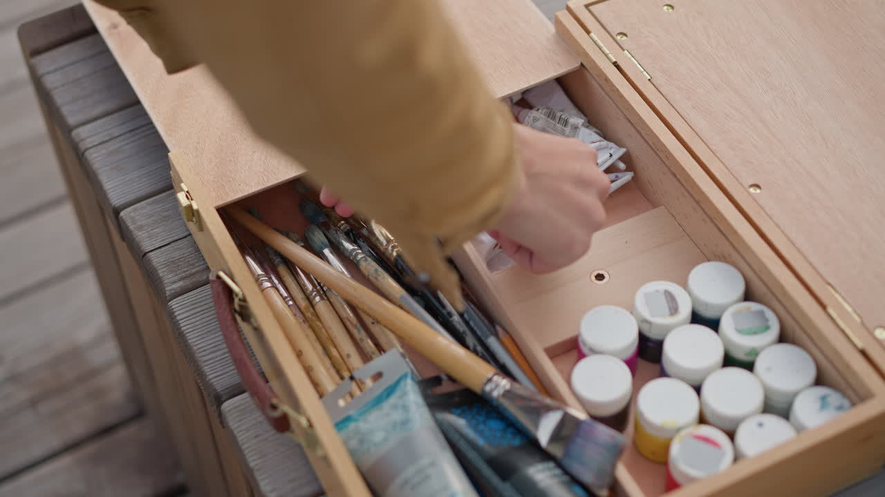 Um artista prepara a caixa de tinta e a paleta de madeira, organiza os pincéis e os tubos em um deck desgastado, close-up das mãos selecionando pincéis, apertando a tinta, colocando potes e misturando cores para um estúdio relaxante