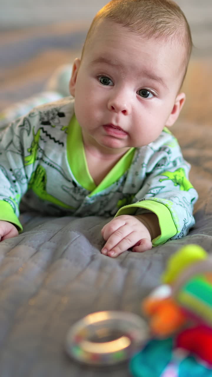 Lovely beautiful kid lies on big bed and looks at a bright toy in front of him. Grey background in blur. Vertical video