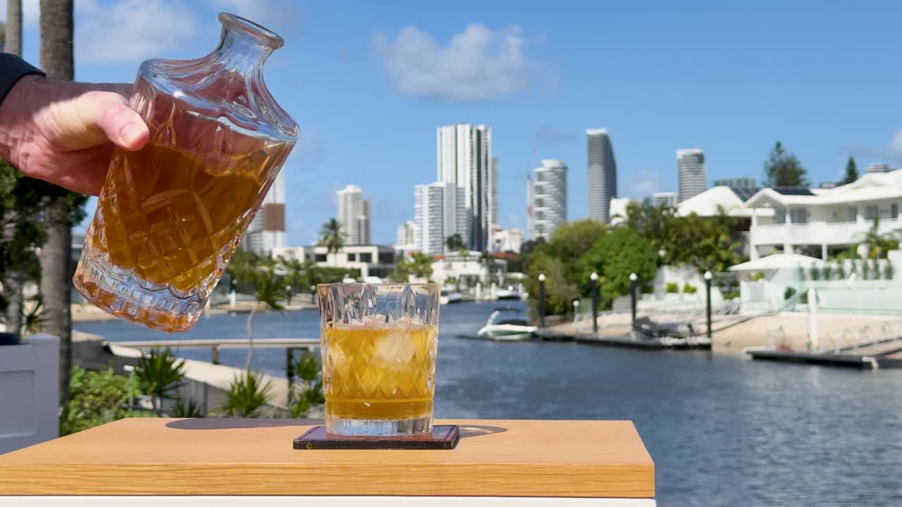 A hand pours whiskey from a glass decanter into an ice-filled tumbler on a wooden table, set against a sunny waterfront cityscape