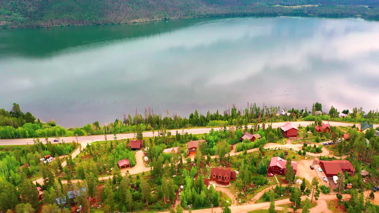 vista aérea de un pequeño pueblo de montaña al lado de un lago con el reflejo de nubes y colinas con coches conduciendo por la carretera viajando más allá de las casas de la aldea a lo largo de la costa de un embalse de montaña rocosa