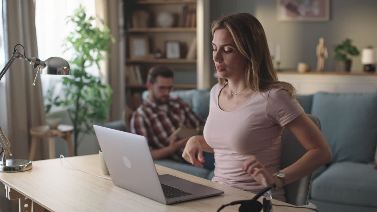 Woman working on laptop at home with man reading in background