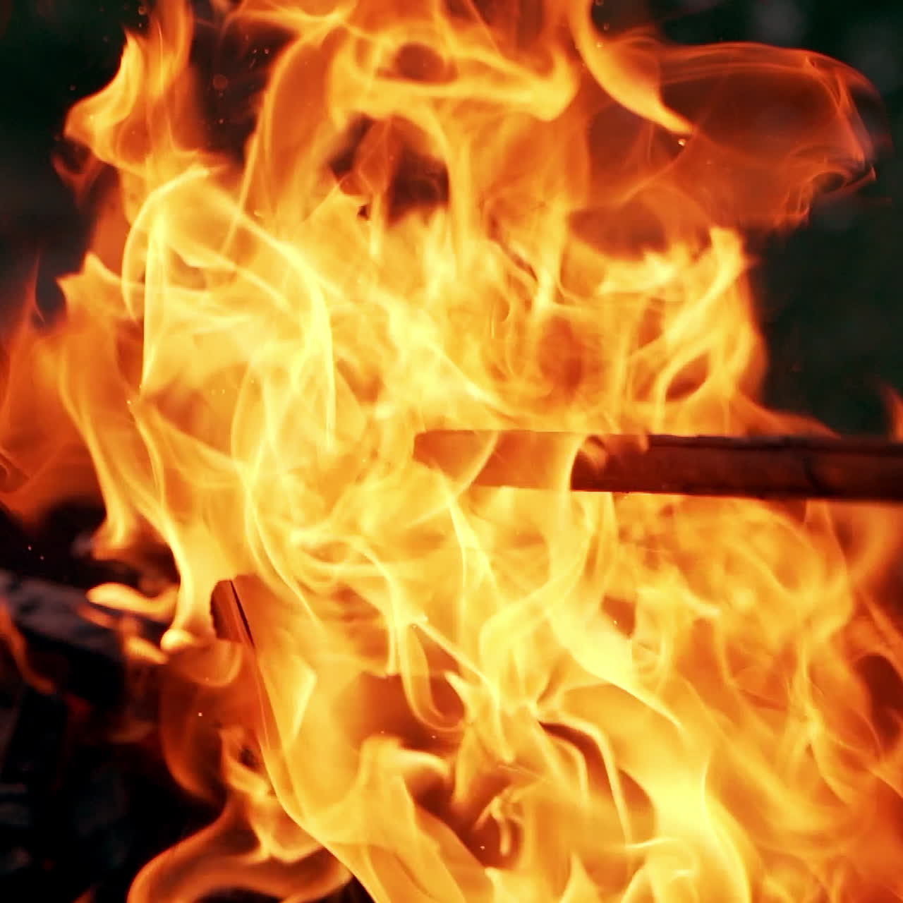 Wooden stick is mixing logs in bright fire. Orange flame from burning firewood outdoors. Smoldered logs in vivid fire flame.