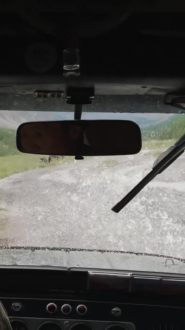 POV dall'interno di un veicolo fuoristrada che percorre una strada di montagna fangosa nella Valle di Akturu, Repubblica dell'Altai, Russia, con gocce di pioggia sul parabrezza e tergicristallo in movimento.