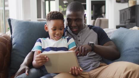 Video-of-happy-african-american-father-and-son-sitting-on-sofa-and-using-tablet