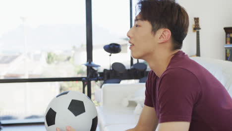 happy asian male teenager watching tv with football and sitting in living room