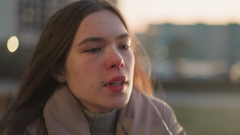 a close-up shot of a young woman wearing a peach jacket, with a contemplative expression on her face, lost in deep thought. the blurred urban background enhances the focus on her introspective mood