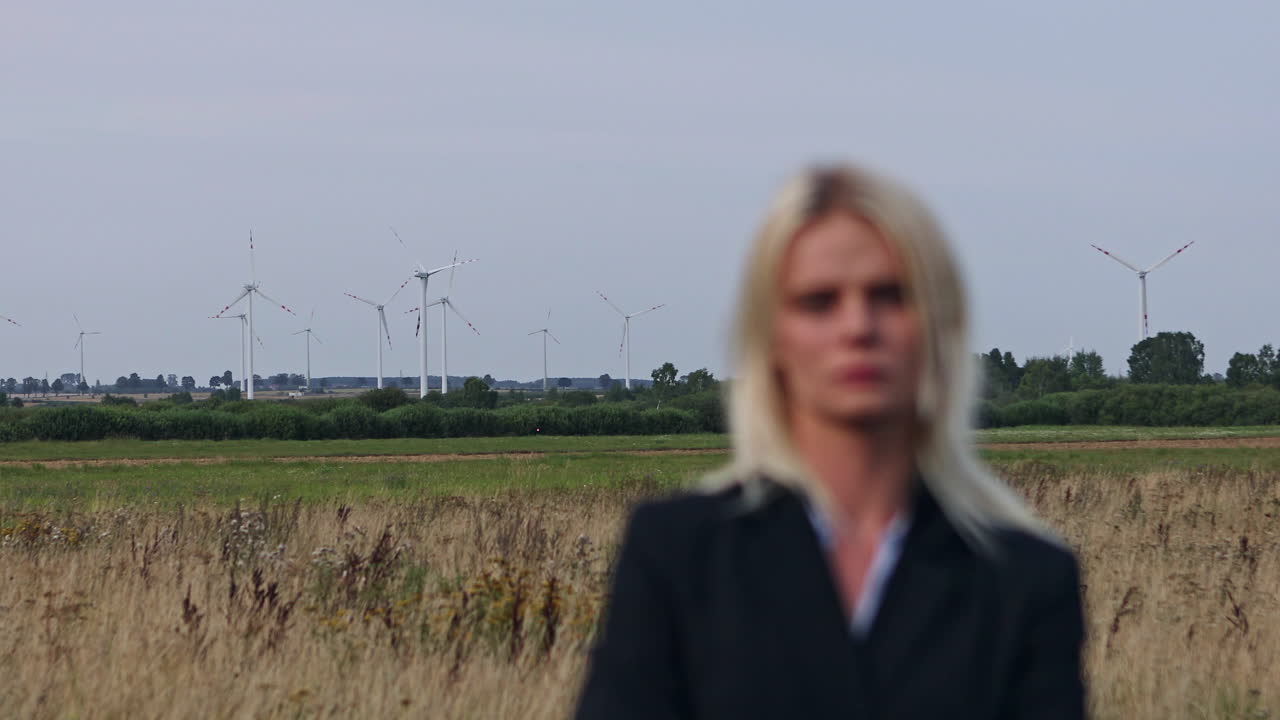 Clean energy from wind turbines. Focus shifts from the young businesswoman standing in the field to the wind turbines behind her. Clean green energy production technology