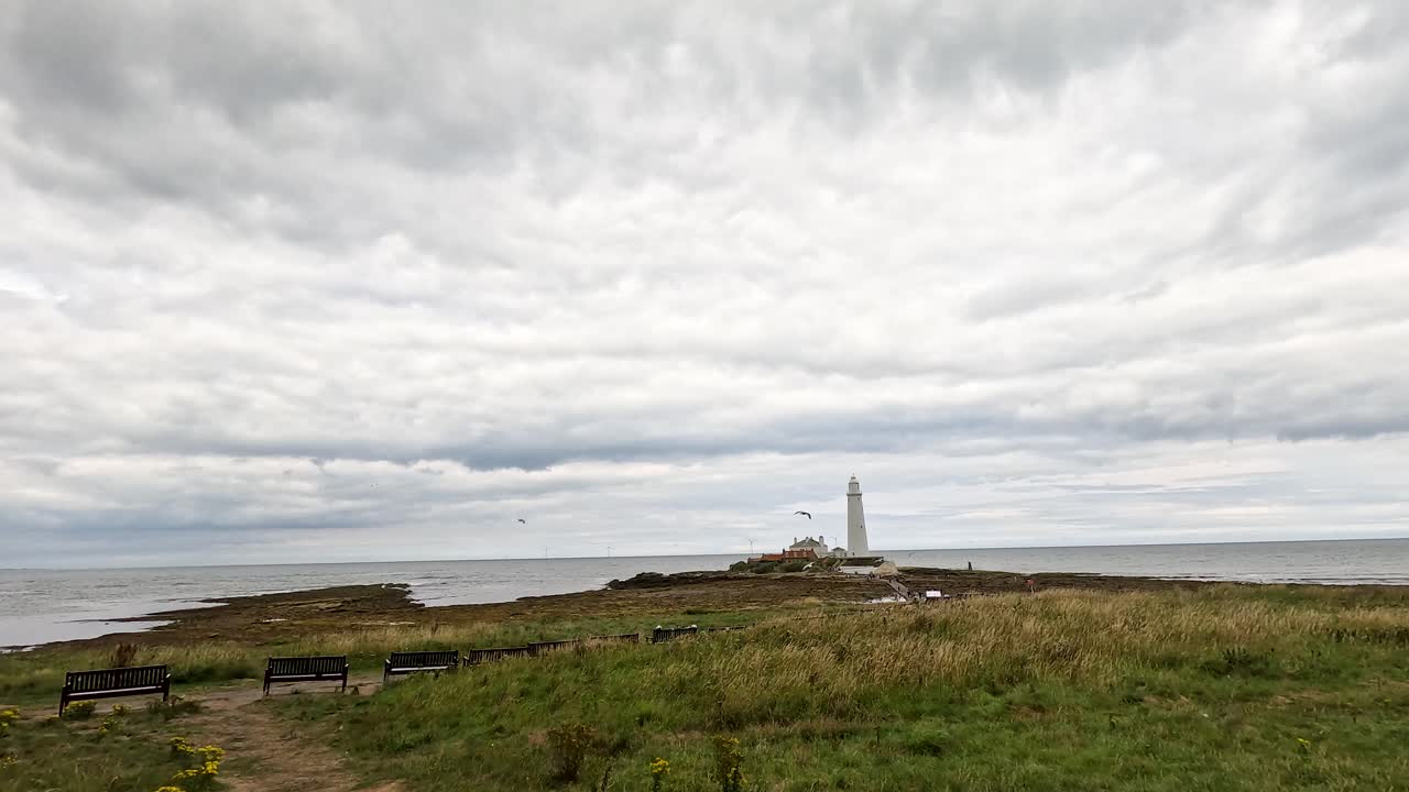 A wide, overcast coastal landscape slowly pans right, unveiling a distant white lighthouse on a rocky shoreline with benches and grassy foreground