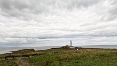 a wide, overcast coastal landscape slowly pans right, unveiling a distant white lighthouse on a rocky shoreline with benches and grassy foreground