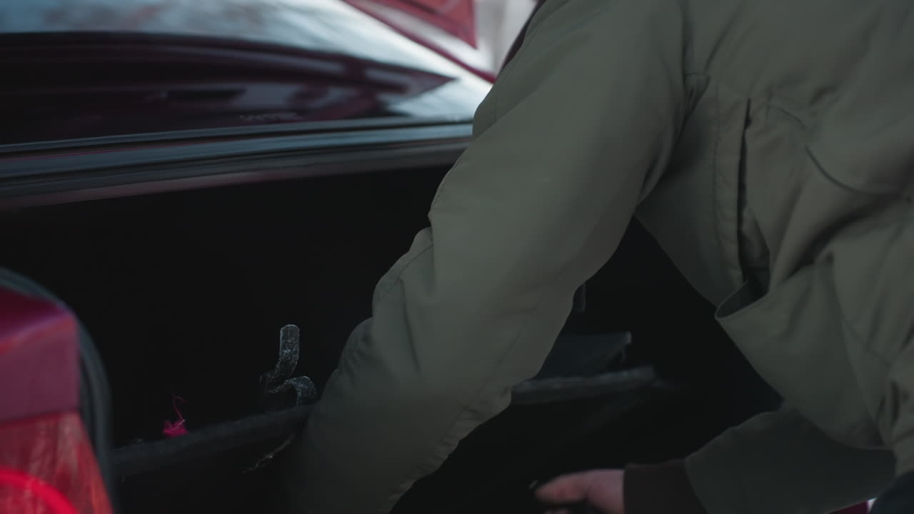 Side view of person in green winter jacket reaching into car boot to remove jack tool, trunk lid open, snowy ground and soft natural lighting in background