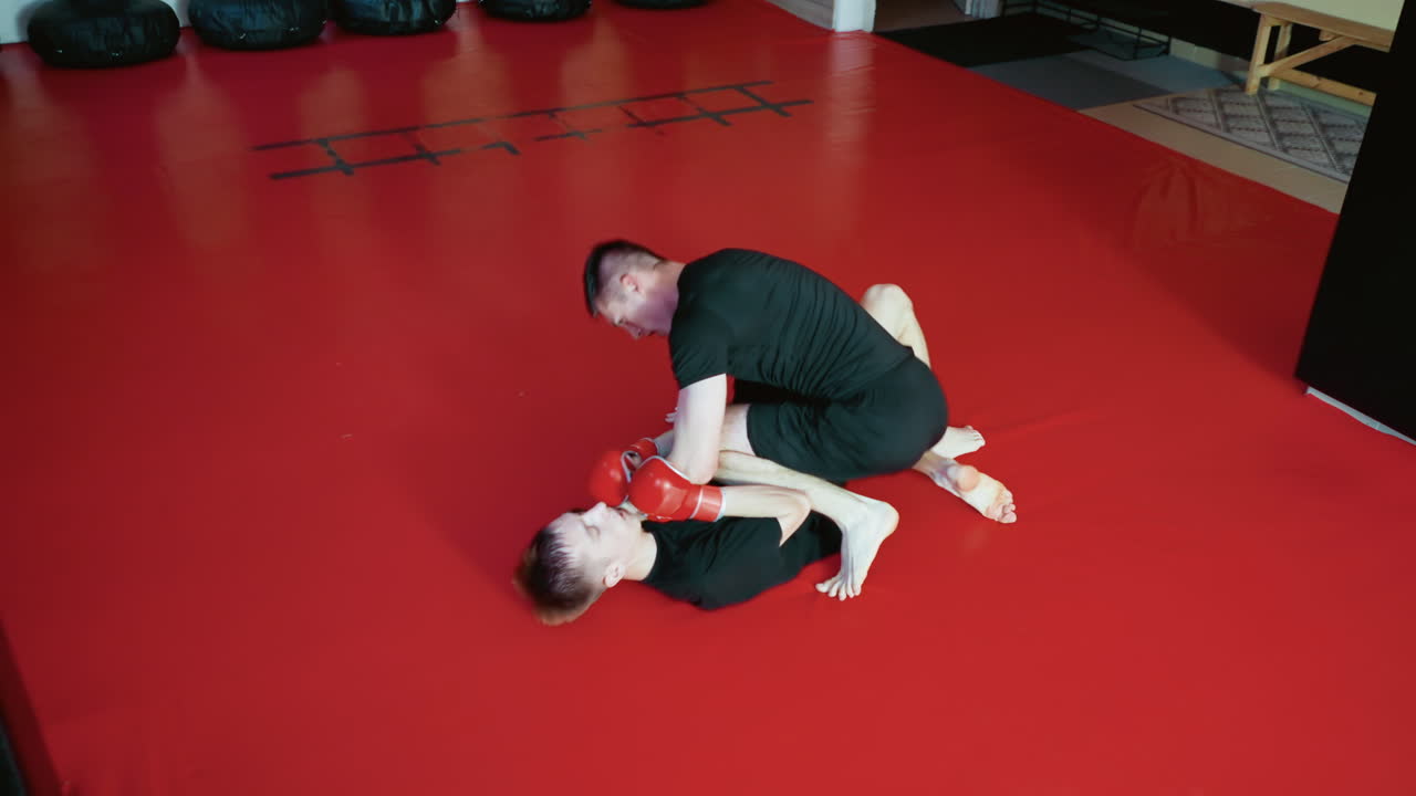Wrestlers grappling during sparring inside gym, wearing gloves and black outfits, performing combat takedown move with strength and technique on red mat floor surrounded by punching bags