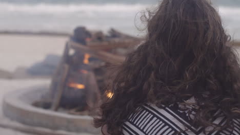 Brunette-woman-in-sweater-enjoys-sitting-next-to-the-fire-pit-by-the-ocean-side