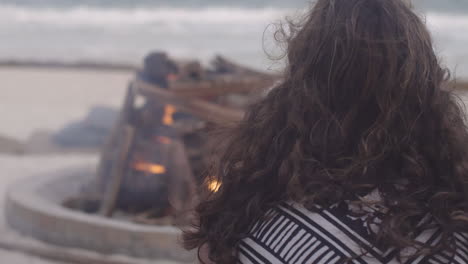 brunette woman in sweater enjoys sitting next to the fire pit by the ocean side