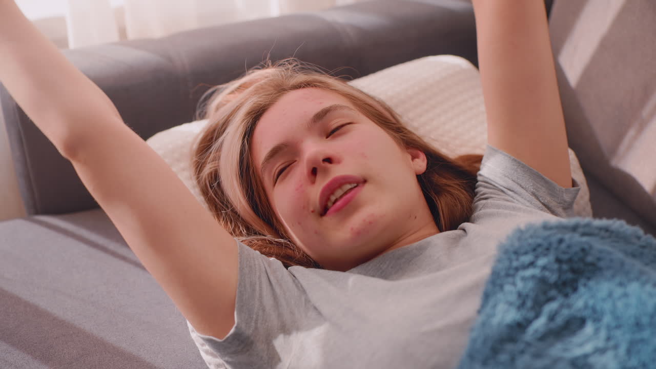 Close up of girl waking up with excitement stretching on couch, lying on white pillow with soft blanket, sunlight streaming through curtains creating peaceful calm atmosphere of morning comfort