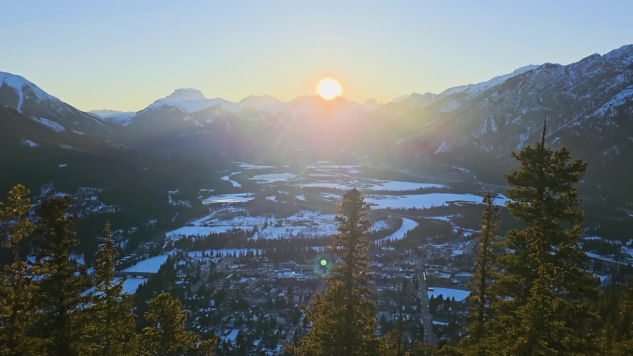 Phong cảnh rộng lớn của Banff, Alberta vào mùa đông, với mặt trời đang lặn trên Dãy núi Rocky của Canada, thung lũng phủ đầy tuyết, những hàng thông, đỉnh núi, thị trấn thơ mộng bên dưới và khung cảnh du lịch hoang sơ.