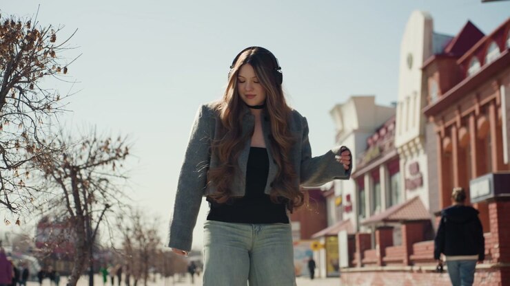 Bella donna asiatica che cammina sul marciapiede della città, studentessa casual durante il tragitto di mezzogiorno oltre negozi di mattoni rossi, pantaloni larghi e cappotto, passo sicuro e rilassato, luce solare soffusa e lunghe ombre, ritratto spontaneo di strada.
