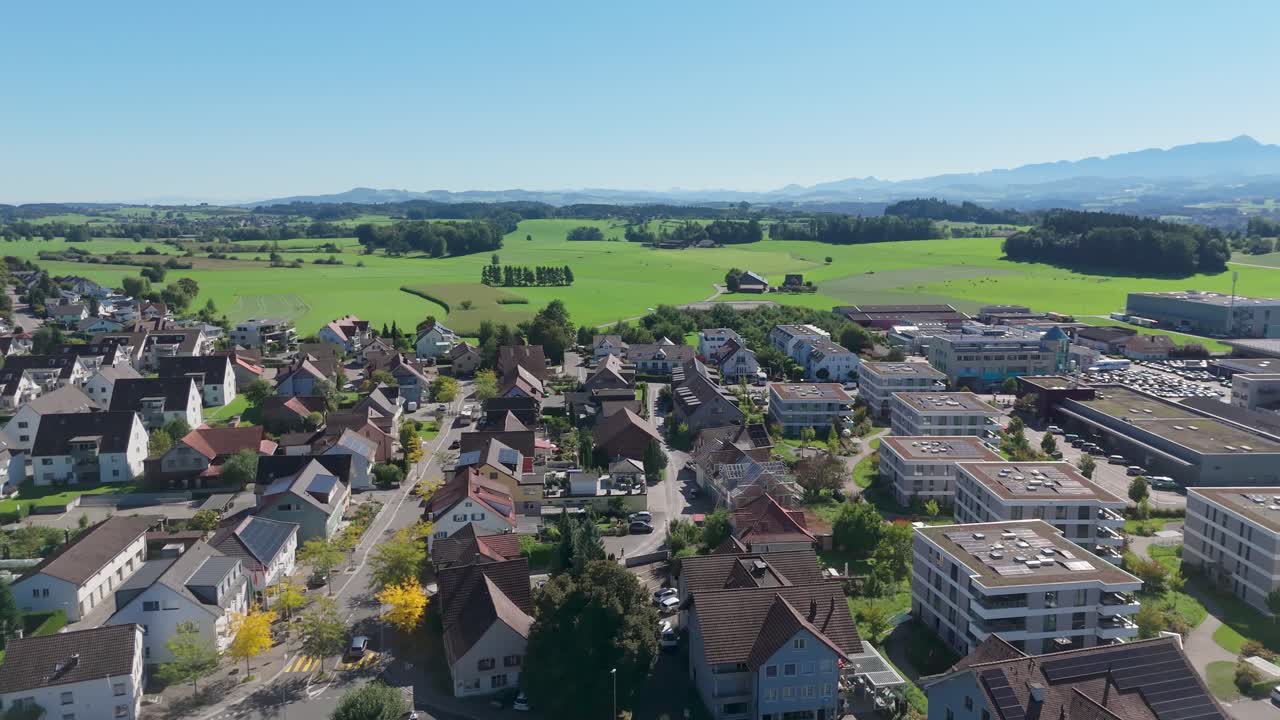 Modern Swiss town neighborhood with solar panels on houses. Rural farm fields in background at sunny summer day. City of Zuzwil in Switzerland with alps in distance. Aerial dolly wide