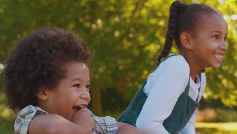 Two-Smiling-Children-Climbing-On-Fence-On-Walk-In-Summer-Countryside