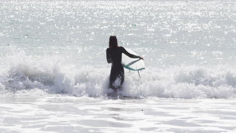 Vista-Trasera-De-Una-Mujer-De-Raza-Mixta-Caminando-Hacia-El-Mar-Llevando-Una-Tabla-De-Surf