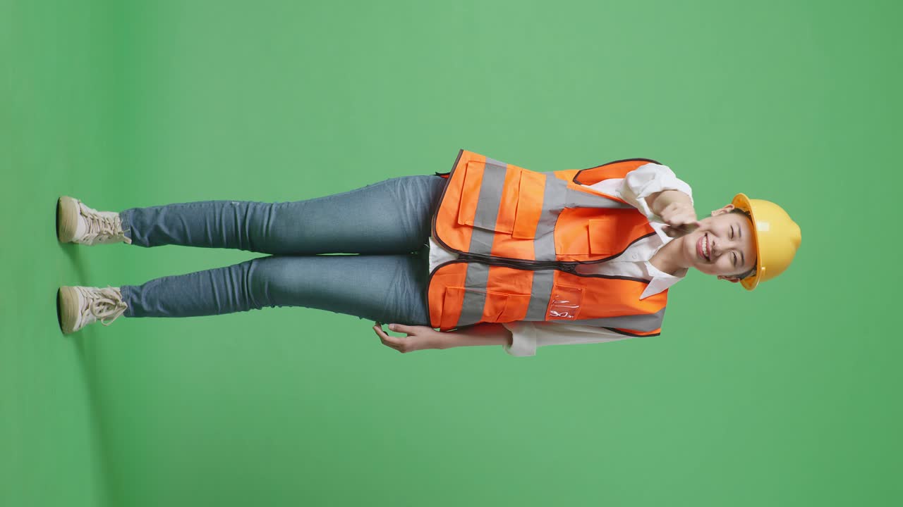 안전 헬을 입은 아시아 여성 엔지니어가 웃으며 가을 만지며 녹색 스크린 배경 스튜디오에 서있는 동안 당신을 가리키고 있습니다.