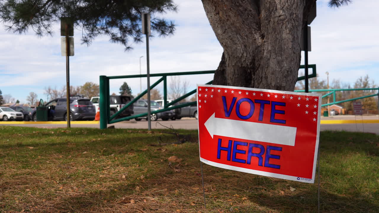 Vote Here Sign Pointing Left with Person Sitting in Car in Parking Lot Background