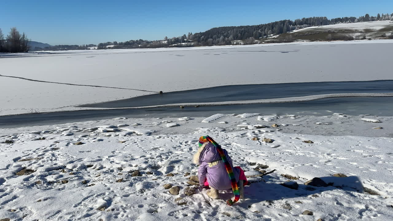 A child dressed in colorful winter clothing plays near the frozen Rottachsee, with snow-covered ground and a partially frozen lake stretching into the distance under a clear blue sky.