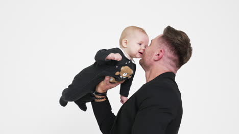 strong man holding his infant son with one hand. cute child in bodysuit with teddy smiles to dad adorably. white backdrop.