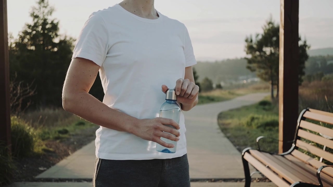 Donna in maglietta bianca che si allunga per svitare il tappo di metallo su una bottiglia trasparente d'acqua su un sentiero del parco, vicino a una panchina.