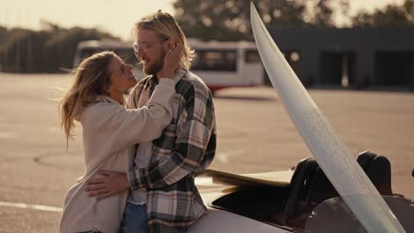 A-blonde-girl-in-a-white-sweatshirt-stands-and-hugs-her-blonde-boyfriend-with-a-beard-in-a-checkered-shirt-near-a-white-convertible-with-a-white-surfboard-on-a-large-square-in-the-city