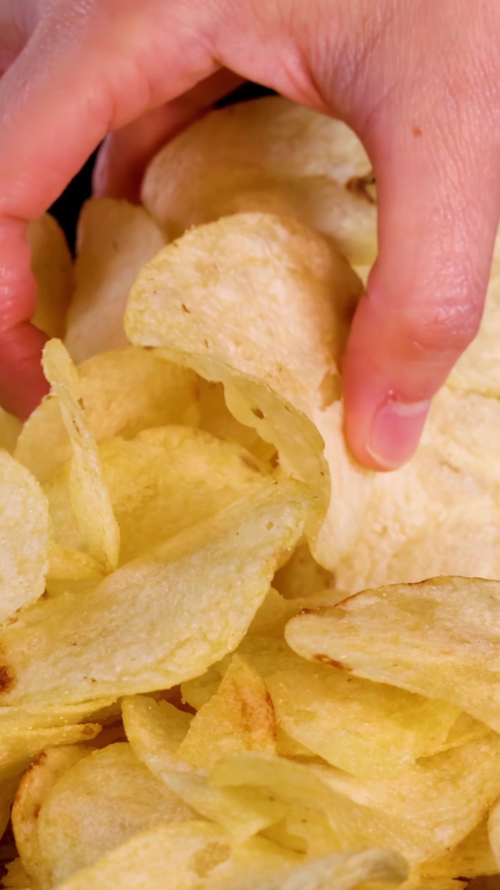 Human hand reaches into pile of potato chips, grabbing snack under bright studio lighting, close-up