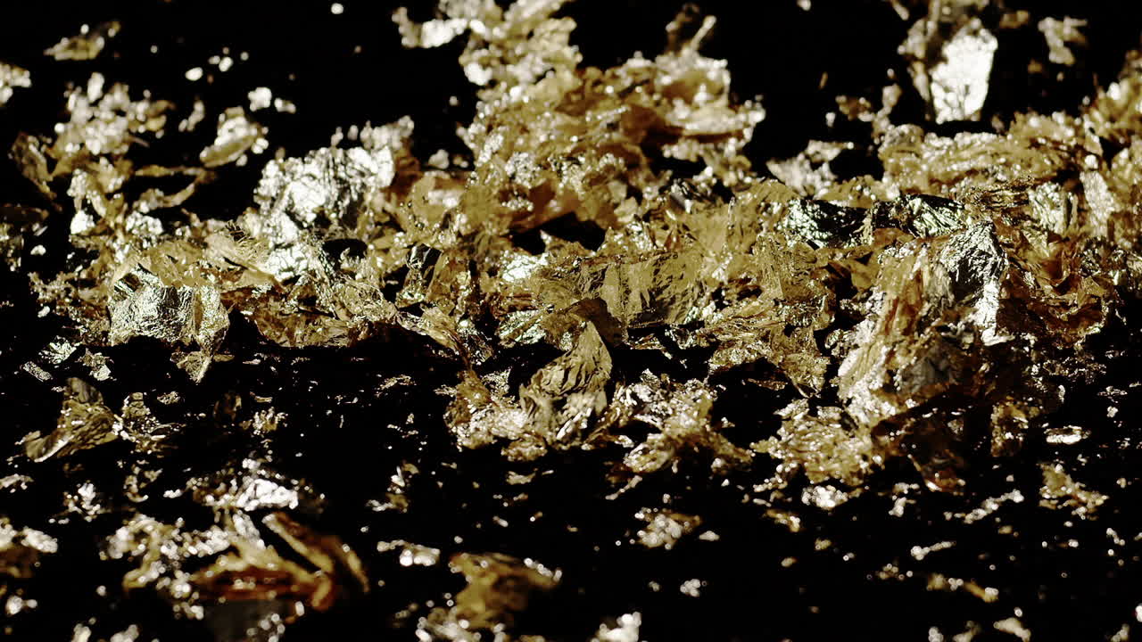 Extreme slow motion close-up of chunky gold flakes blown in a blast of wind from the right side of the screen.