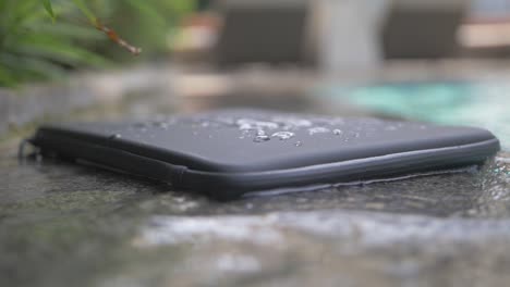 detail shot of a black, basic, waterproof laptop case with lines engraved in it lies on the side of a pool with splashes and drops of water on it